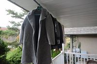 Grey wool coat hanging on a hanger in an outdoor covered porch area, showing large collar and front pockets.