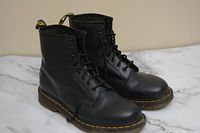 Pair of black Dr. Martens AirWair leather boots shown from front side angle, showing classic eight eyelet lace-up, yellow stitching, rubber sole.
