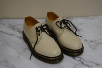 Pair of off-white leather Dr. Martens shoes with black laces displayed on a marble surface.