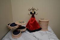 Red dress jewelry holder with metal hooks alongside two beige hand-painted floral spinning table jewelry holders with multiple tiers and compartments.