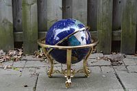 Side angle view of the semi-precious stone globe on brass tripod stand sitting outdoors on paving stones with wooden fence backdrop.