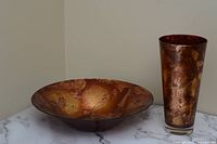 Glass bowl and vase set featuring copper hue coloration placed on marble surface against beige wall.