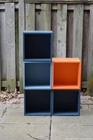 Photo shows 5 Ikea Eket storage boxes in black, blue and orange colors stacked outdoors against a wooden fence. Boxes have no doors or hardware, gently used condition.