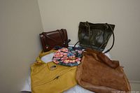 A collection of five handbags including a yellow bag, a brown leather bag, dark green bag, small floral painted bag, and another brown handbag all displayed on a white cloth against a beige wall.