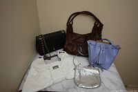 Lot of five purses arranged on a marble surface including the black quilted purse, brown shoulder bag, lavender bucket purse, white J.Crew purse, and silver shimmery purse