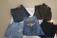 Five pairs of jeans in varying denim washes, folded and arranged on a flat surface showing variety in brands and styles.