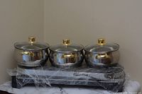 Photo of three polished stainless steel pots with glass lids and gold-tone handles, arranged on a black electric heating base wrapped in plastic.