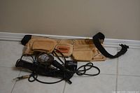 Photo showing brown leather Mastercraft utility tool belt with multiple pouches and an adjustable black strap lying on the floor. In front are several utility lights with black handles and attached cords.