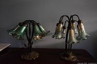 Two tulip-shaped lamps with greenish glass shades on a dark surface against a dark background, showing the heavy bronze bases and curved stems. One lamp is taller with six shades, the other shorter with five, one shade missing.