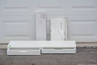 White Ikea Alex desktop or shelf unit with two drawers placed on the pavement in front of a garage door, showing signs of use.