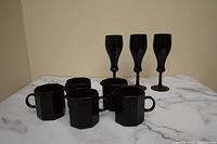 Photo of three black octagonal mugs with handles, and three black stemmed glasses arranged on a white marble surface.