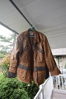 Brown leather coat with multiple zippered and button flap pockets, hanging on green hanger outdoors. Lightly worn condition.