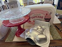 Photo showing red cake stand with tag, glass cake stand, cupcake storage container, and 3D bear cake pan.
