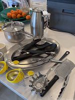Photo showing assorted kitchen utensils including melon ballers, pizza cutter, tongs, metal cheese grater, pasta fork, and stainless steel items on a counter.