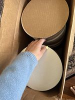 Hand holding a circular white cake board inside a cardboard box filled with similar boards, showing thickness and size (8 inches).