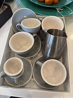 Top view of coffee cups (espresso, cappuccino, latte) with matching saucers and stainless steel milk frother arranged in a white tray on a table.