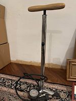 Full view of the floor bicycle pump showing wooden handle, stainless steel body, pressure gauge, black hose, and foot pedal on patterned rug.