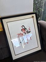 Framed print showing the full artwork of a stylized robin perched on abstract branches with leaves in red and black colors. The print is under glass in a black frame with a white mat. A gold numbered medallion 73 is visible on the mat corner.