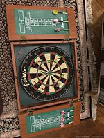 Opened wooden dartboard cabinet showing dartboard with numerous darts inserted and chalk scoreboards on doors.