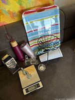 Overview of all kitchen items displayed on a couch, showing a striped tray, water bottle, fruit basket, grater, scale, vinegar/olive dispensers, roll holder, towel, and candle holder.