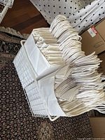 Photo showing many white paper gift bags with twisted handles neatly arranged inside a white wire basket with a fabric liner on a patterned rug.