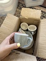 Photo of six light grey ceramic espresso cups and matching saucers packed in a cardboard box, showing the cups stacked inside the saucers.