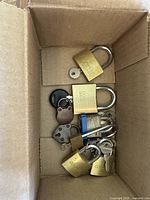 Photo showing various locks inside a cardboard box including brass padlocks marked GRK, small vintage locks, and keys.