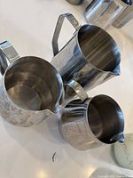 Three stainless steel milk frother jugs arranged on a white surface showing different sizes and spouts for pouring.