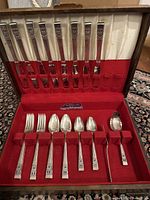 Full flatware set displayed open in original box showing knives, forks, spoons, butter spreader and serving spoon held in red velvet compartments with decorative silver handles.