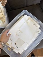 Stack of white flattened cardboard bakery boxes with die-cut plastic window, held in hand over a plastic container.