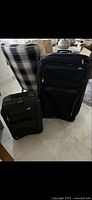 Photo showing two black Samsonite suitcases posed on floor near a plaid chair. The sizes are visually distinct with one extra large and one carry-on size suitcase. Both suitcases are upright, displaying multiple zippered compartments and fabric texture.