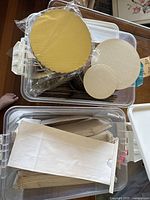 Plastic bin containing stacks of round mini cake boards with gold tops and white bottoms in two different sizes, alongside a large stack of white paper bags.