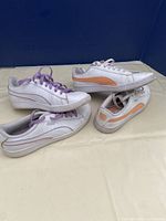 Two pairs of Puma sneakers displayed showing side and top views, highlighting the purple and orange color accents.