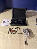 Front view of the black iPad Mini case with integrated Bluetooth keyboard, USB charging cable, mini key fob camera in package, and instruction manual on a beige surface with a blue background.