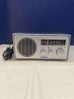 Front view of the silver Sangean radio showing speaker, control knobs, and tuning scale