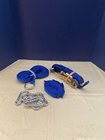 Set of blue heavy duty tie down straps, one with ratchet, two with carabiners, and four loose metal carabiners on flat surface