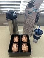Oggi Pumpmaster insulated thermos shown next to original box, four copper clad hammered Moscow mule mugs in black insert box, and one clear plastic iced beverage cup with blue lid and straw.