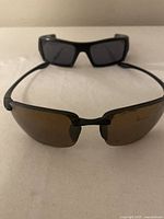 Two pairs of sunglasses: one Oakley with black frame and dark lenses, one Maui Jim with black frame and brown lenses. Minor scratches and discoloration visible on lenses.