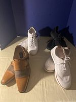 Photo showing four pairs of men's shoes: one tan and beige monk strap dress shoes, one white pair of low-top sneakers, one white lace-up sneakers, and one black lace-up sneakers.