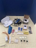 Photo showing all items in the lot including the phone sanitizer, Oster razor, cords, bottle toppers, fairy lights and other small items arranged on a beige fabric background.