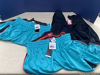 Three pieces of clothing laid out: two green basketball shorts and one black track pant on a beige surface with blue background.