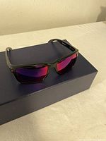 Front view of the Oakley sunglasses resting on a dark blue box, showing red tinted lenses and black frame.