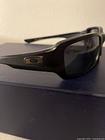 Close-up of black frame Oakley sunglasses showing left temple with Oakley logo metal badge and tinted lens