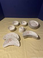 Top view showing 4 floral Limoges teacups, 6 saucers stacked, and 6 crescent-shaped bone plates laid out on beige cloth under direct light