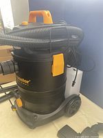 Full view of the wet/dry vacuum showing black body with yellow handle and latches, wheels and coiled hose on top.