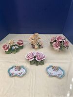 Five porcelain items arranged on cream cloth against a blue background, showing Capo di Monte rose figurines and Meiko rose decorative plates.