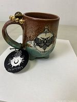 Side view of ceramic mug showing brown and green glaze, moth print, and 22k gold beads with tigers eye crystal on handle.