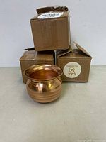 Three cardboard boxes stacked behind one copper lota with polished surface and wide flared rim
