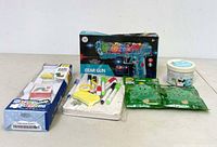 Lot of kids games and toys including Story Dream machine, gear gun box, face paint kit, container, and two green foil bags