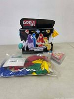Boxed 'Bop It!' kids game with packaging showing game instructions and colorful characters. Next to it is a sealed bag containing a variety of colorful balloons.